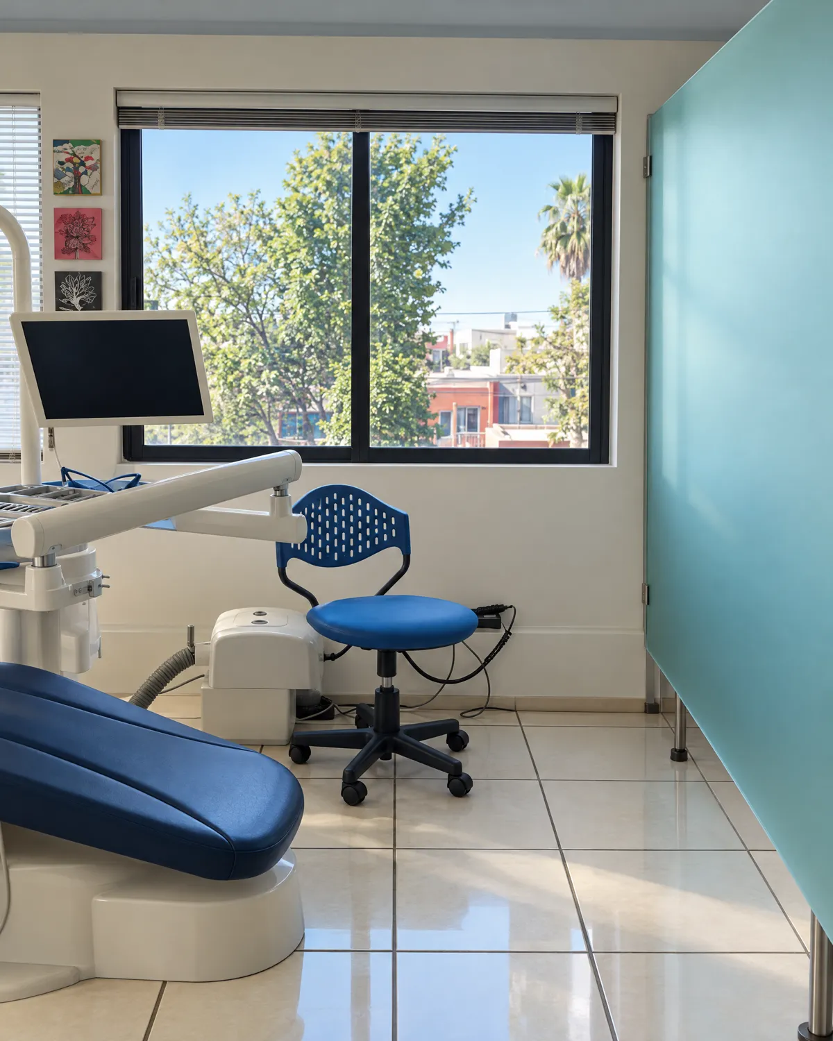 Área clínica: sillón dental azul, banco y ventana con árboles, junto a una mampara de vidrio turquesa
